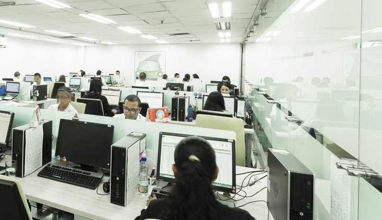 Espacio de oficina con varios empleados trabajando en estaciones de computadoras, con escritorios modernos y mamparas de vidrio.