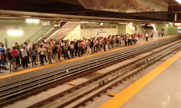 Multitud de pasajeros esperando en un andén del metro con dos vías vacías visibles y líneas de seguridad amarillas en el borde del andén.