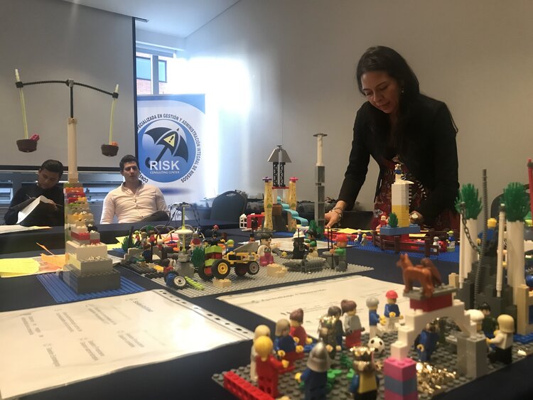 Una mujer interactúa con una exhibición de un modelo de Lego durante un taller, con los participantes y los carteles del taller al fondo.