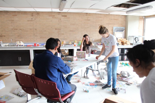 Personas participando en un taller de artesanía en una mesa con materiales de arte, dentro de una habitación con paredes de ladrillo y una gran ventana.