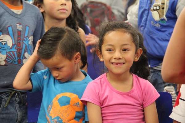 Una joven sonriendo a la cámara con una camisa rosa, sentada junto a un niño con una camisa azul que mira hacia abajo, entre un grupo de niños en un salón de clases.