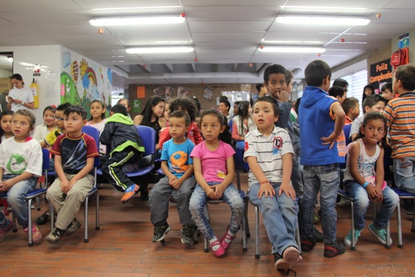 Niños sentados y de pie en un aula colorida, atentos y con carteles educativos en las paredes.