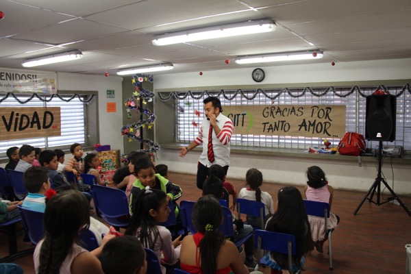 Un hombre con una camisa a rayas entretiene a un salón de clases de niños, con adornos y un cartel que dice "gracias por tanto amor" al fondo.