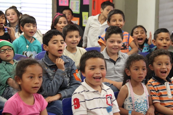 Grupo de niños sentados atentamente con expresiones emocionadas en un salón de clases, algunos mirando hacia el frente.