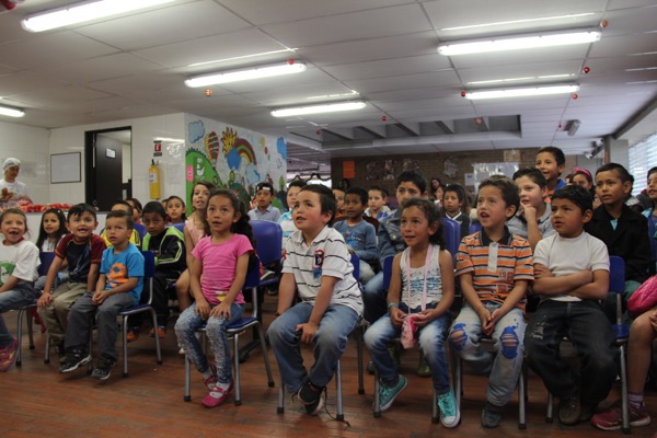 Grupo de escolares sentados y de pie en un aula, mirando atentos y ligeramente hacia arriba, con coloridas obras de arte en las paredes.