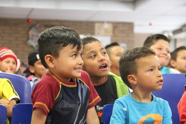 Niños pequeños sentados en fila en un salón de clases, mirando hacia arriba con expresiones de interés y entusiasmo.