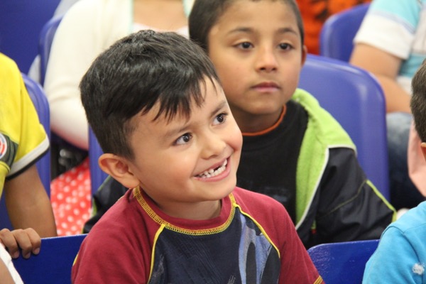 Un niño de pelo corto sonriendo ampliamente entre la multitud, mirando a su izquierda, con otros niños al fondo.