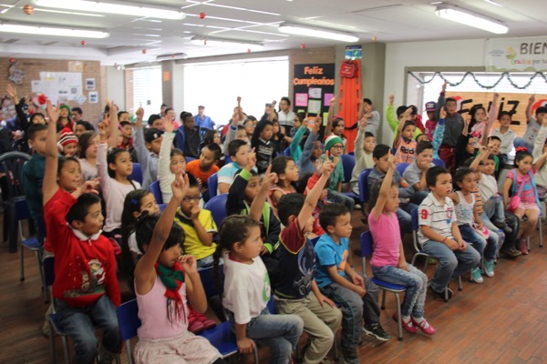 Niños levantando la mano en un aula decorada para una celebración, con adultos de pie al fondo.