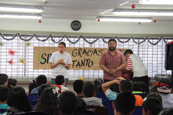 Una escena de aula con dos adultos hablando ante un grupo de niños sentados; una pancarta que dice "sin gracias tanto" cuelga al fondo.