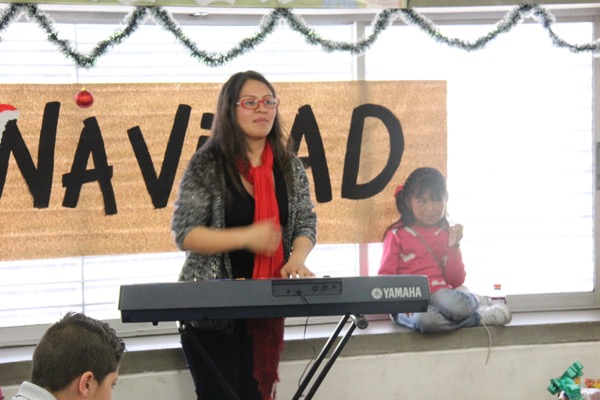 Una mujer toca un teclado electrónico en un evento navideño mientras una niña sentada cerca, con adornos de fondo.