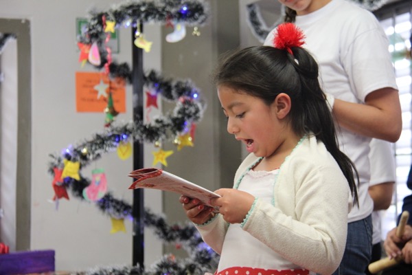 Una niña leyendo en voz alta un periódico en una habitación decorada durante un evento festivo, con adornos navideños de fondo.