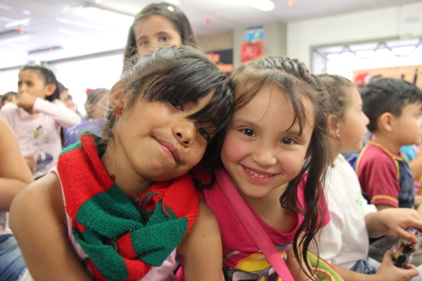 Dos niñas sonriendo y abrazándose, una de ellas con un pañuelo rojo y verde, en un aula llena de otros niños.