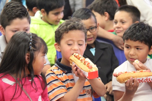 Un niño muerde un hot dog rodeado de otros niños en una animada reunión.