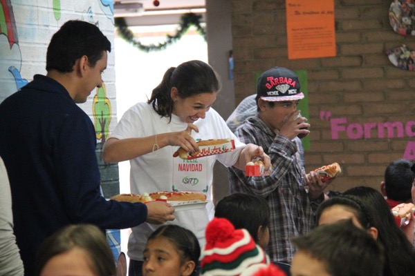 Adultos sirviendo pizza a niños en una sala decorada festivamente durante un evento navideño.