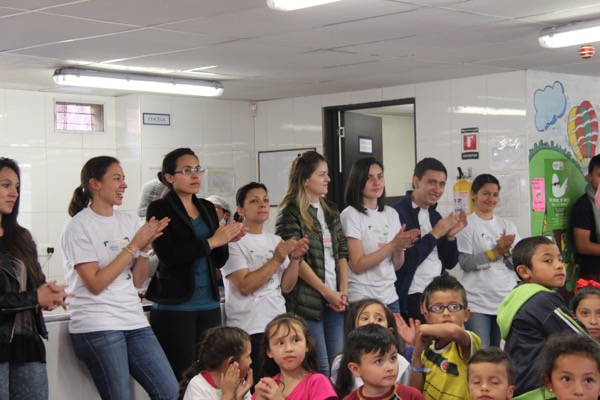 Grupo de adultos y niños aplaudiendo en una habitación muy iluminada con paredes blancas y decoraciones coloridas.