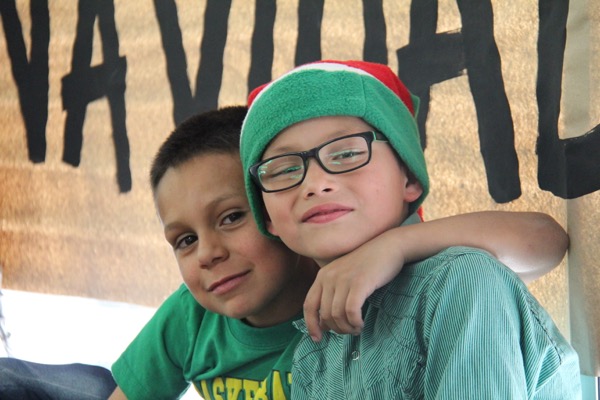 Dos jóvenes sonriendo a la cámara, uno con gafas y un gorro verde, sentados juntos bajo un cartel con el texto "navital".