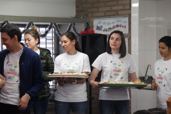 Grupo de adultos con camisetas de "feliz navidad" y diademas de astas sirviendo comida en un evento festivo en una cocina.