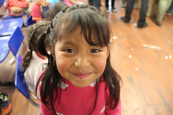 Niña de cabello oscuro con un suéter rosa sonriendo a la cámara en un salón de clases con otros niños al fondo.