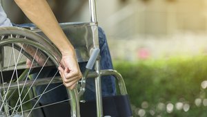 Primer plano de la mano de una persona aferrada al volante de una silla de ruedas al aire libre, con un fondo verde borroso, que representa PcDOV: Personas con Discapacidad de Origen Vial.