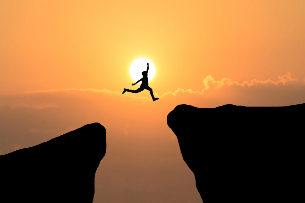 Una silueta de una persona saltando entre dos bordes de acantilados contra un vibrante fondo de puesta de sol.