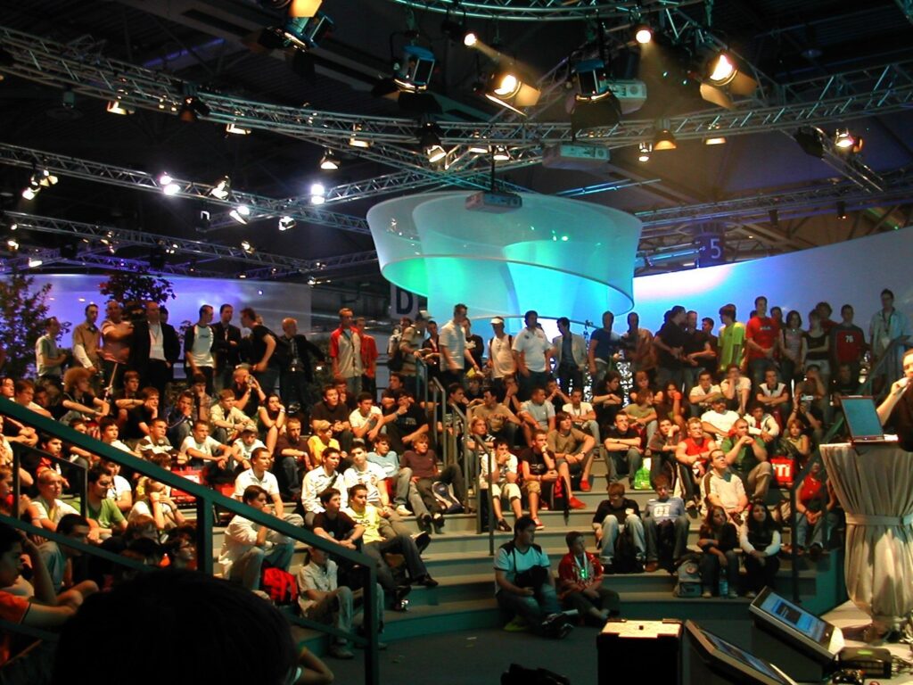Un evento tecnológico lleno de gente con gente sentada y de pie en una sala iluminada por luces azules y verdes, centrándose en un escenario de presentación.