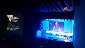 Una pantalla grande que muestra a una oradora en una conferencia en un auditorio oscuro, con "gobierno del estado de Victoria Melbourne" visible.