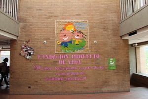 Interior de un edificio con un mural de dos niños de dibujos animados en una pared de ladrillos, acompañado de un texto inspirador en español sobre la protección de la vida en ciudad bolívar.