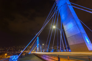 Puente colgante iluminado por la noche con luces azules y soportes imponentes, con vistas a un paisaje urbano.