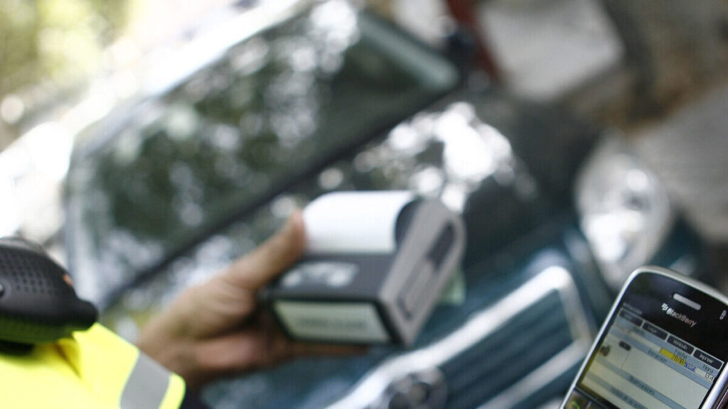 Primer plano de la mano de un oficial de control de estacionamiento usando un dispositivo digital para escanear la matrícula de un automóvil, con un teléfono inteligente también visible.